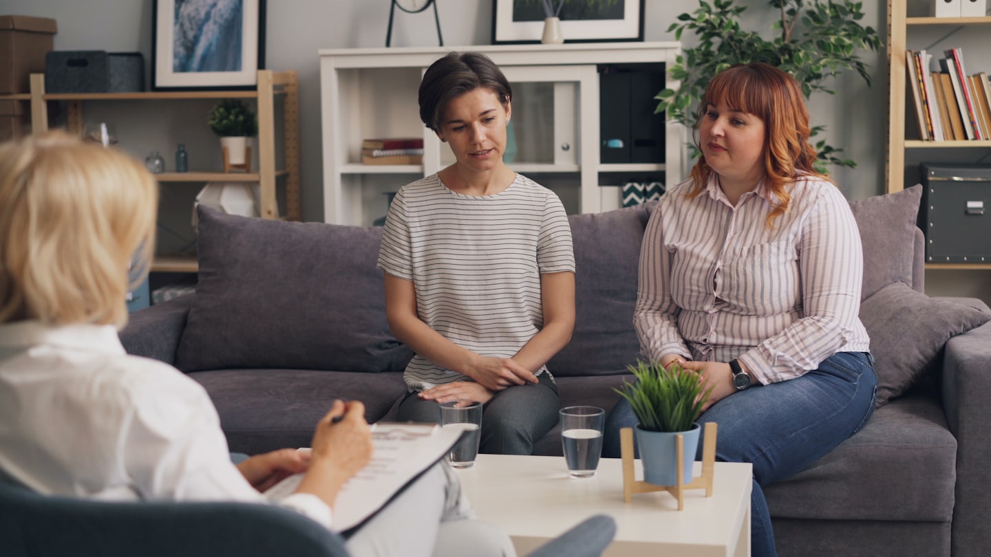Therapeutin auf Klemmbrett berät zwei Frauen auf dem Sofa.